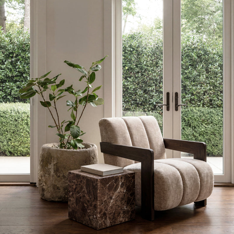 A modern upholstered armchair with a tufted back and seat, dark wood arms, and a low marble side table in front, set in a bright room with tall windows, greenery outside, and a potted plant.