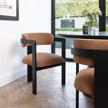A modern dining chair with a warm tan upholstered seat and curved backrest, paired with a black metal frame, shown in a bright dining room setting next to a dark wood table.