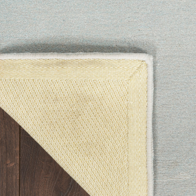 Close-up of a cream-colored woven rug corner with a white border, shown partially folded over a dark wood floor against a light gray background.