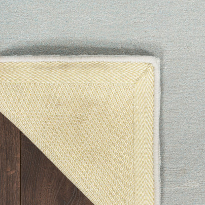 Close-up of a cream-colored woven rug corner with a white border, shown partially folded over a dark wood floor against a light gray background.