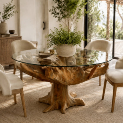A round glass-top dining table with a natural wood root-style base, shown in a bright dining room with cream upholstered chairs and potted plants.