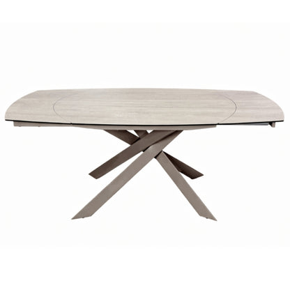A modern oval dining table with a light beige wood-grain top and a unique X-shaped base in a taupe-brown finish, shown on a plain white background.