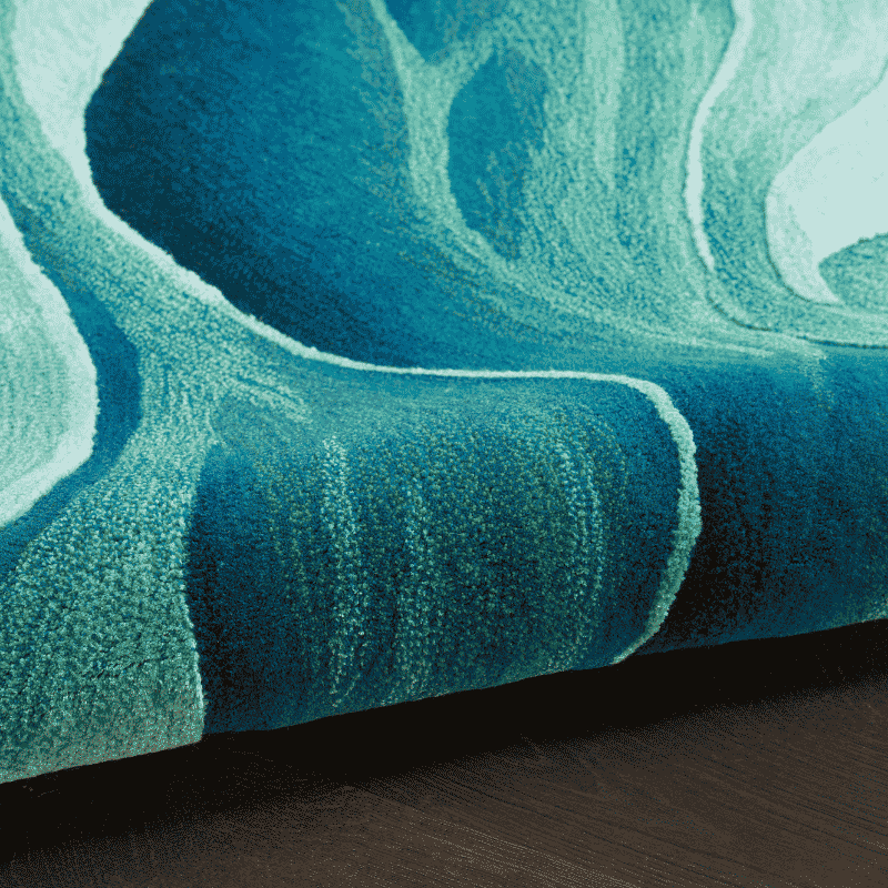A close-up of a plush area rug with an abstract, fluid wave pattern in shades of teal, turquoise, and deep blue, shown folded over to reveal its texture and a dark wood floor underneath.