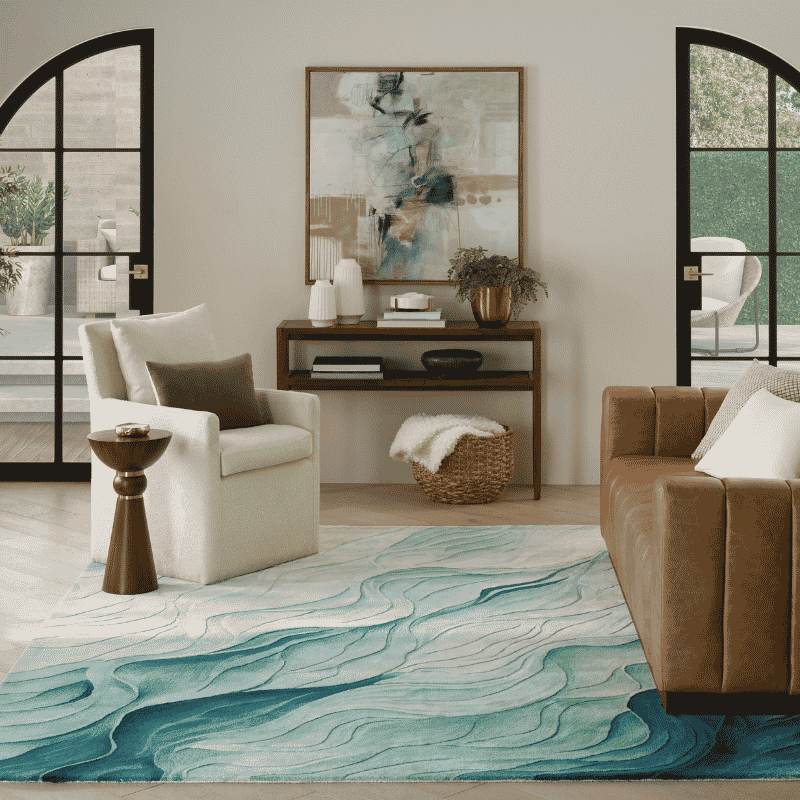 A large area rug with a wavy, fluid abstract pattern in shades of teal, blue, and white, styled in a modern living room setting with a brown leather sofa, a white armchair, and a wooden console table.