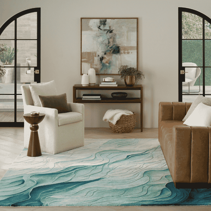 A large area rug with a wavy, fluid abstract pattern in shades of teal, blue, and white, styled in a modern living room setting with a brown leather sofa, a white armchair, and a wooden console table.