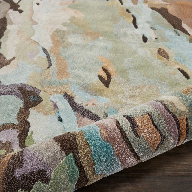 A close-up of a plush area rug with an abstract, painterly pattern in muted tones of beige, sage green, teal, brown, and lavender, shown folded over to reveal its pile and texture.