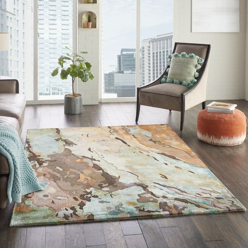 An abstract area rug with a watercolor-like pattern in beige, brown, teal, and cream, styled in a modern living room setting with a brown armchair, orange pouf, and city view through large windows.