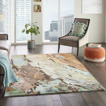 An abstract area rug with a watercolor-like pattern in beige, brown, teal, and cream, styled in a modern living room setting with a brown armchair, orange pouf, and city view through large windows.