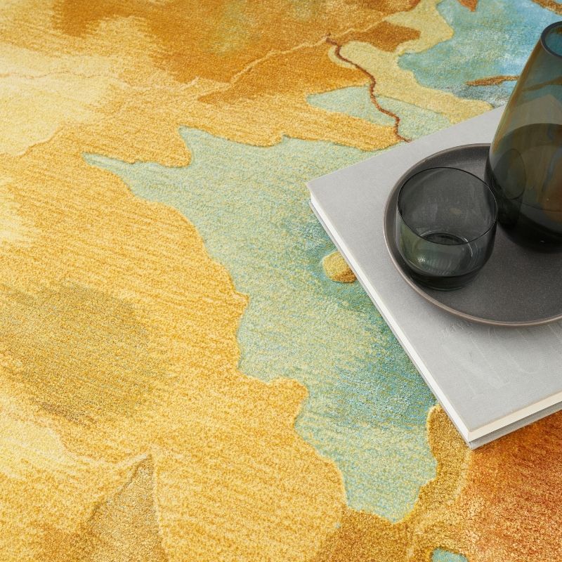 An abstract area rug with a watercolor-like pattern in mustard yellow, beige, light blue, and orange, styled with a gray tray, a dark glass tumbler, and a white book.
