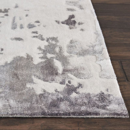 A close-up of a rectangular area rug with a distressed abstract pattern in off-white and gray, shown on a dark brown wood floor.