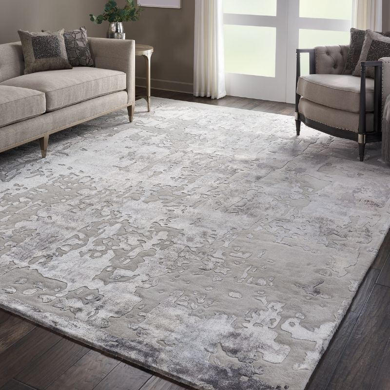 A large rectangular area rug with an abstract distressed pattern in shades of gray, white, and taupe, shown in a modern living room setting with a sofa, armchair, and dark wood flooring.
