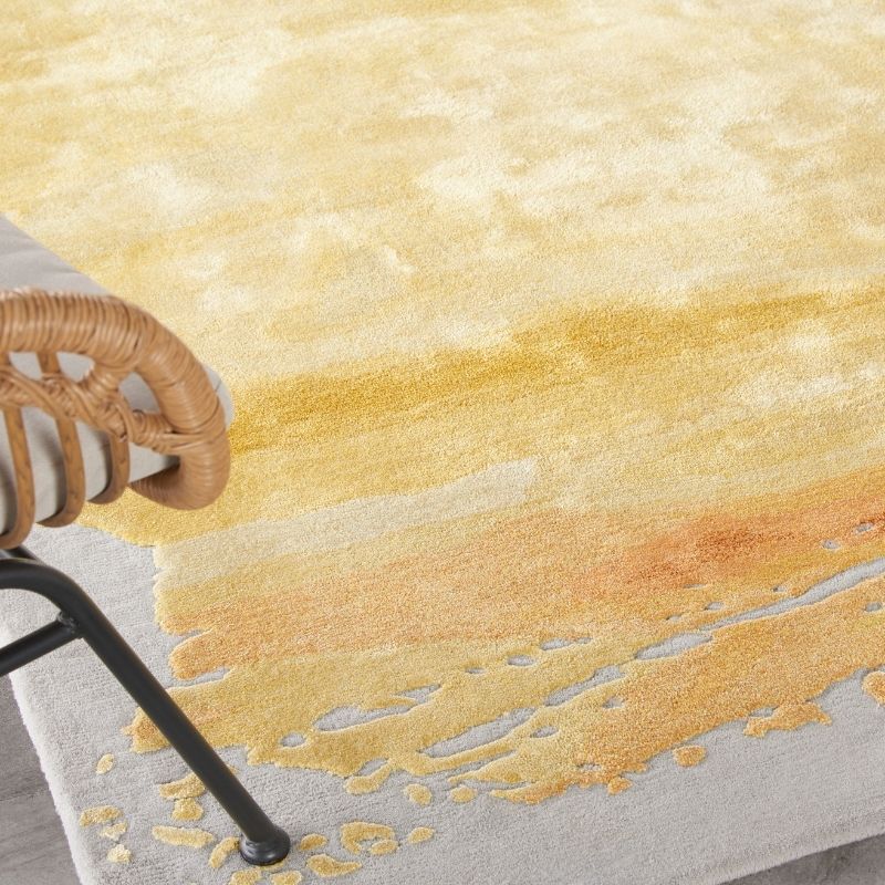 A close-up of a textured area rug with an abstract watercolor design in shades of yellow, orange, and beige on a light gray base. A woven rattan chair is partially visible on the left side.