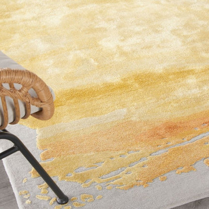 A close-up of a textured area rug with an abstract watercolor design in shades of yellow, orange, and beige on a light gray base. A woven rattan chair is partially visible on the left side.