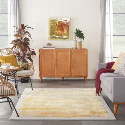 A rectangular area rug with a textured, abstract yellow and beige pattern on a light gray base, styled in a bright, modern living room next to a gray sofa and a wooden credenza.