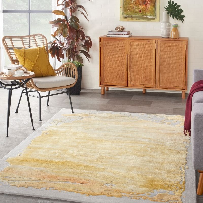 A large rectangular area rug with an abstract watercolor-like pattern in shades of yellow, beige, and soft orange, styled in a bright, modern living room next to a rattan armchair and a wooden credenza.