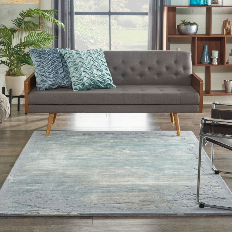 A gray upholstered loveseat with wooden legs sits in a living room setting on a light blue and gray patterned area rug. Two decorative throw pillows in blue and teal are placed on the sofa, with a potted plant, shelving, and window visible in the background.