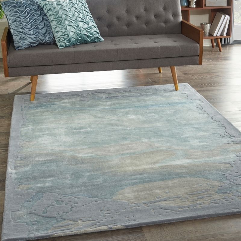 A rectangular area rug with an abstract landscape design in shades of blue, gray, and beige, styled in a living room setting under a gray tufted sofa.