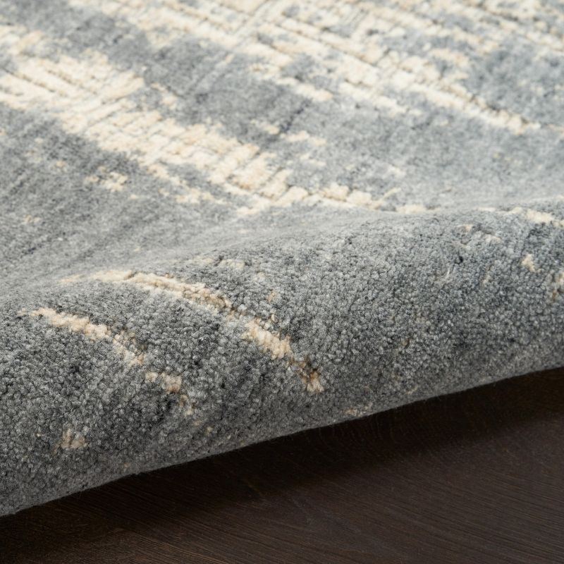Close-up of a textured area rug with a distressed abstract pattern in gray and cream, shown folded over to reveal the pile and underside, resting on a dark wood floor.