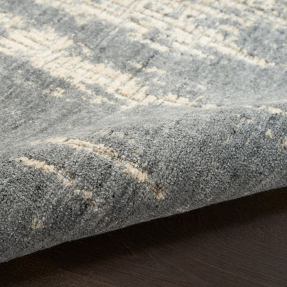Close-up of a textured area rug with a distressed abstract pattern in gray and cream, shown folded over to reveal the pile and underside, resting on a dark wood floor.