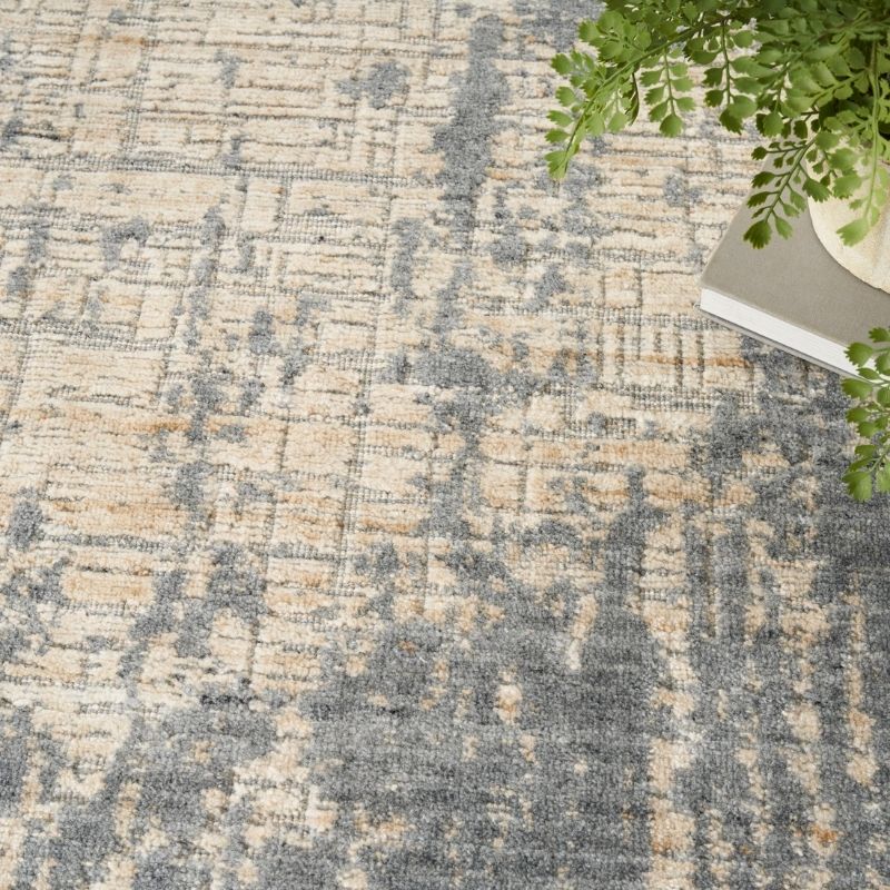 A close-up of a textured area rug with an abstract distressed pattern in beige, gray, and muted blue-gray tones, styled with a green plant and a book in the top right corner.