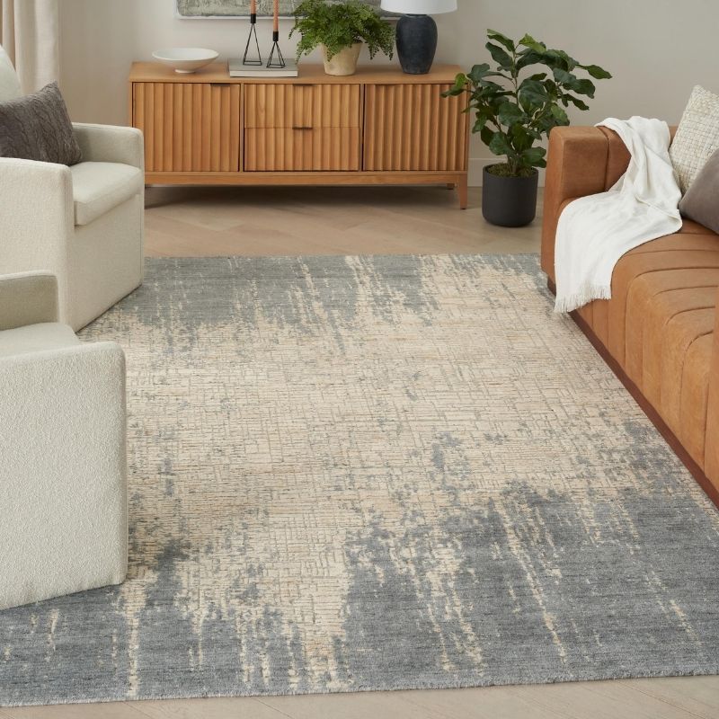 A large rectangular area rug with an abstract distressed pattern in gray and beige, styled in a modern living room setting with a wooden sideboard, armchair, and sofa.