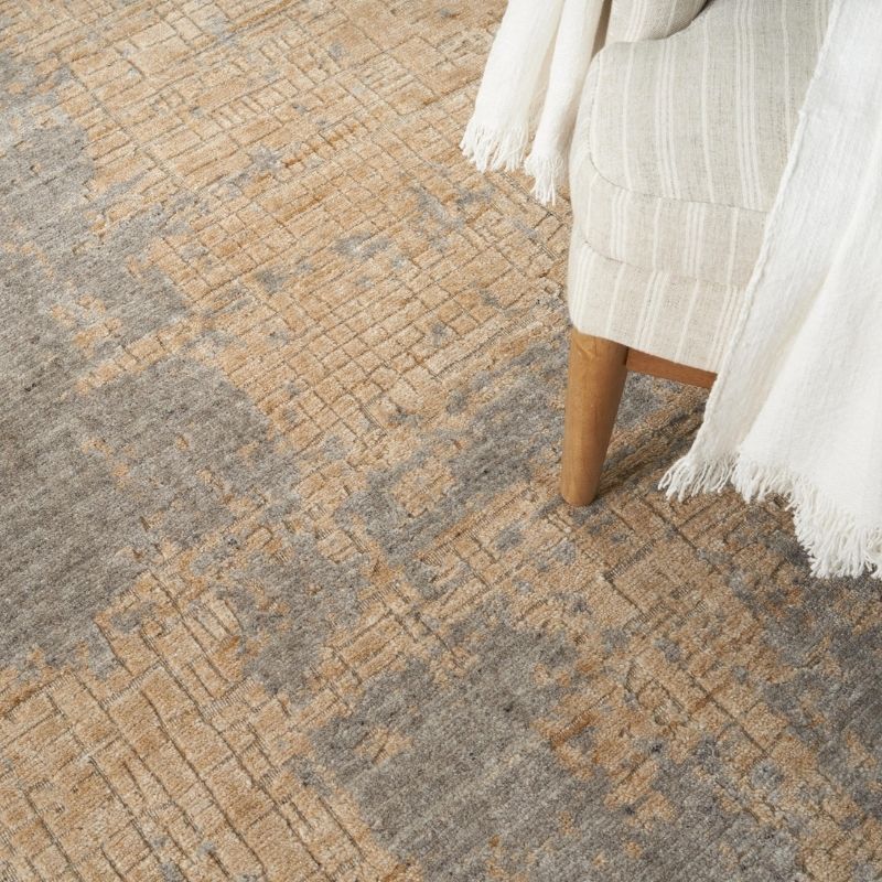 A close-up of a textured area rug with an abstract pattern in beige, tan, and gray, styled with a cream-colored armchair draped with a white fringed throw blanket.