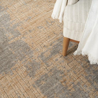 A close-up of a textured area rug with an abstract pattern in beige, tan, and gray, styled with a cream-colored armchair draped with a white fringed throw blanket.