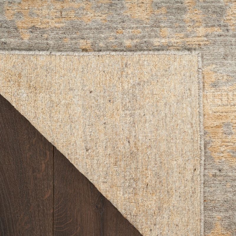 Close-up of a textured area rug corner showing a warm beige central field, a distressed gray and tan outer patterned border, and a dark brown wood surface underneath.