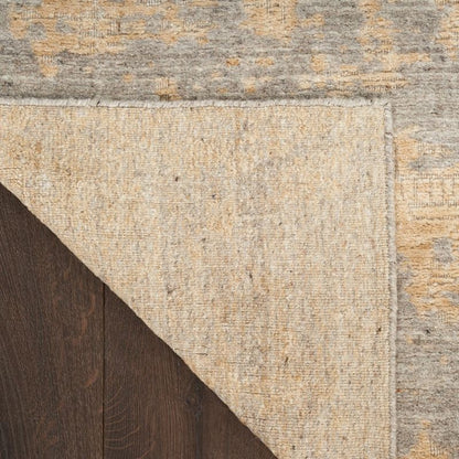 Close-up of a textured area rug corner showing a warm beige central field, a distressed gray and tan outer patterned border, and a dark brown wood surface underneath.