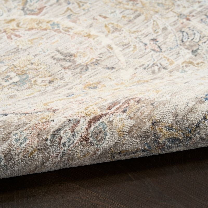 A close-up of a folded area rug showing its detailed traditional pattern in beige, cream, brown, and muted blue tones, resting on a dark wood floor.