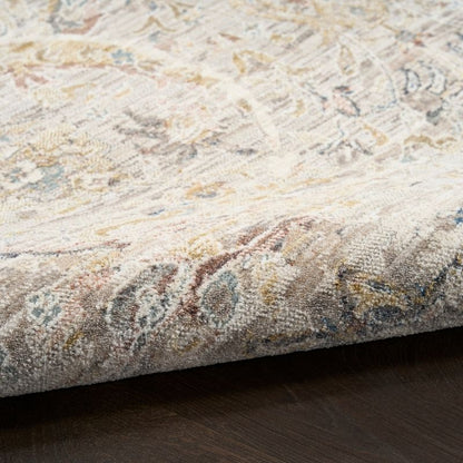 A close-up of a folded area rug showing its detailed traditional pattern in beige, cream, brown, and muted blue tones, resting on a dark wood floor.