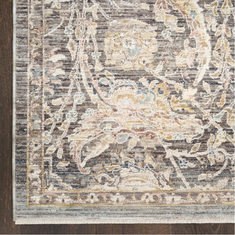 A close-up of a distressed traditional area rug with an ornate floral and vine pattern in muted beige, gray, brown, and soft blue tones, laid over a dark wood floor.