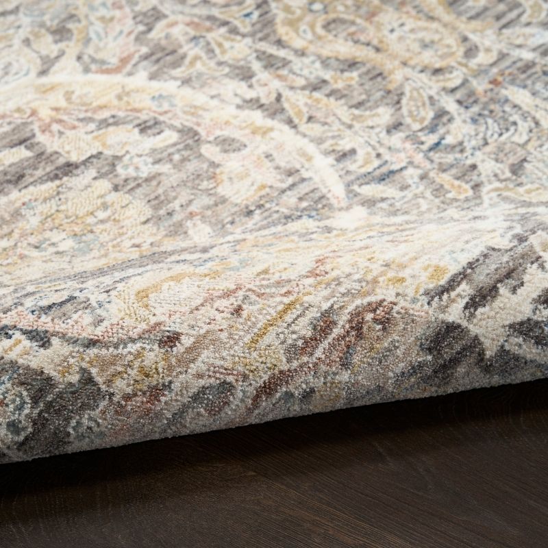 A close-up of a patterned area rug with a traditional paisley design in beige, gray, brown, and muted blue tones, shown folded over to reveal its texture and underside, resting on a dark wood floor.