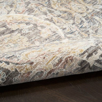 A close-up of a patterned area rug with a traditional paisley design in beige, gray, brown, and muted blue tones, shown folded over to reveal its texture and underside, resting on a dark wood floor.