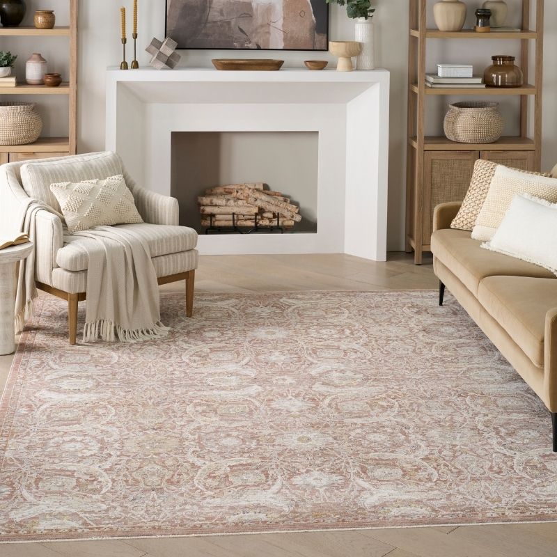 A large rectangular area rug with an ornate traditional floral pattern in dusty blush pink and cream, styled in a bright, modern living room with a beige sofa, striped armchair, and white fireplace.