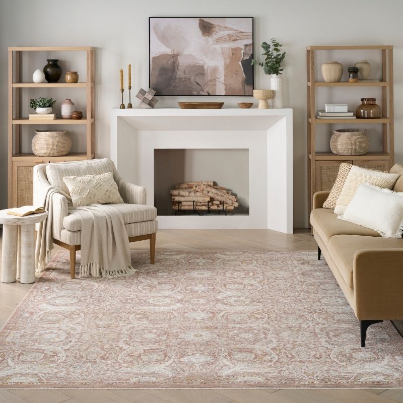A styled living room featuring a large beige and pink ornate area rug on the floor, a cream armchair with a throw blanket, a tan sofa, light wood shelving, and a white fireplace with a framed abstract print.