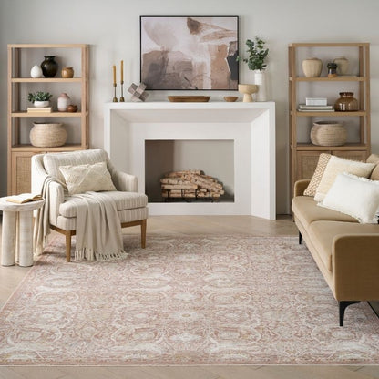 A styled living room featuring a large beige and pink ornate area rug on the floor, a cream armchair with a throw blanket, a tan sofa, light wood shelving, and a white fireplace with a framed abstract print.