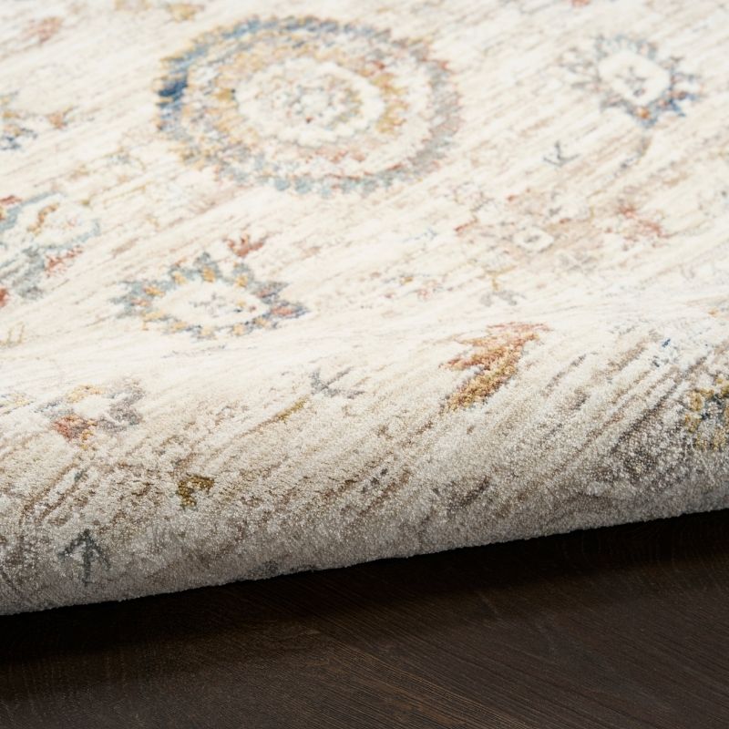 A close-up of a folded area rug showing its textured pile and distressed traditional pattern in cream, beige, muted blue, and brown tones over a dark wood floor.
