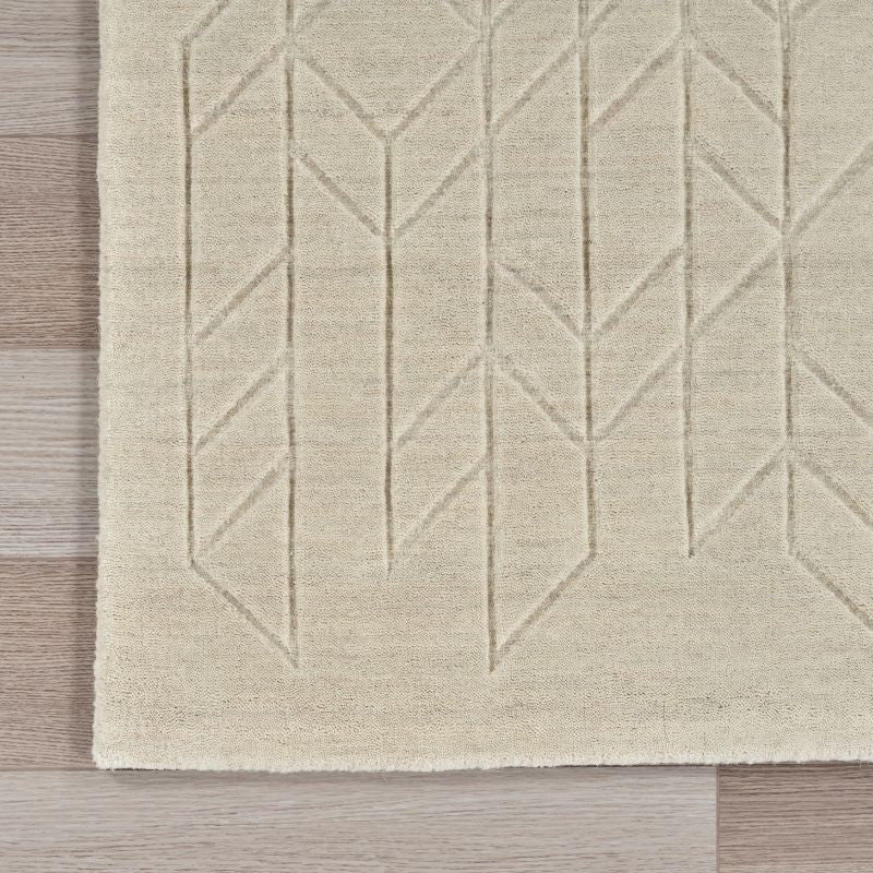 A close-up of a light beige area rug with a geometric, raised chevron-like pattern, shown on a light wood floor.