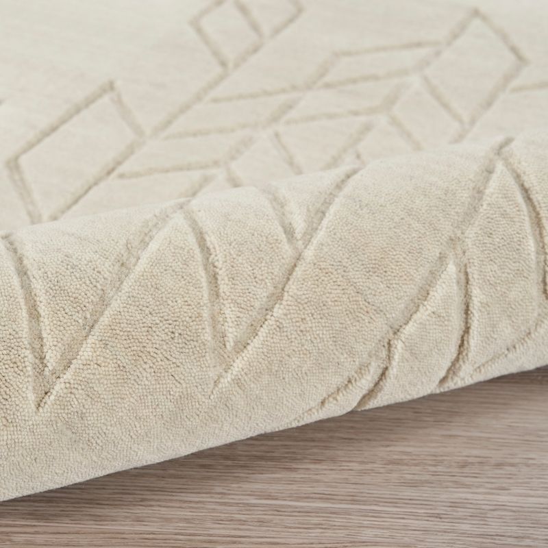 A close-up of a light beige textured rug with a geometric diamond pattern, shown folded over to reveal its pile and texture on a light wood floor.