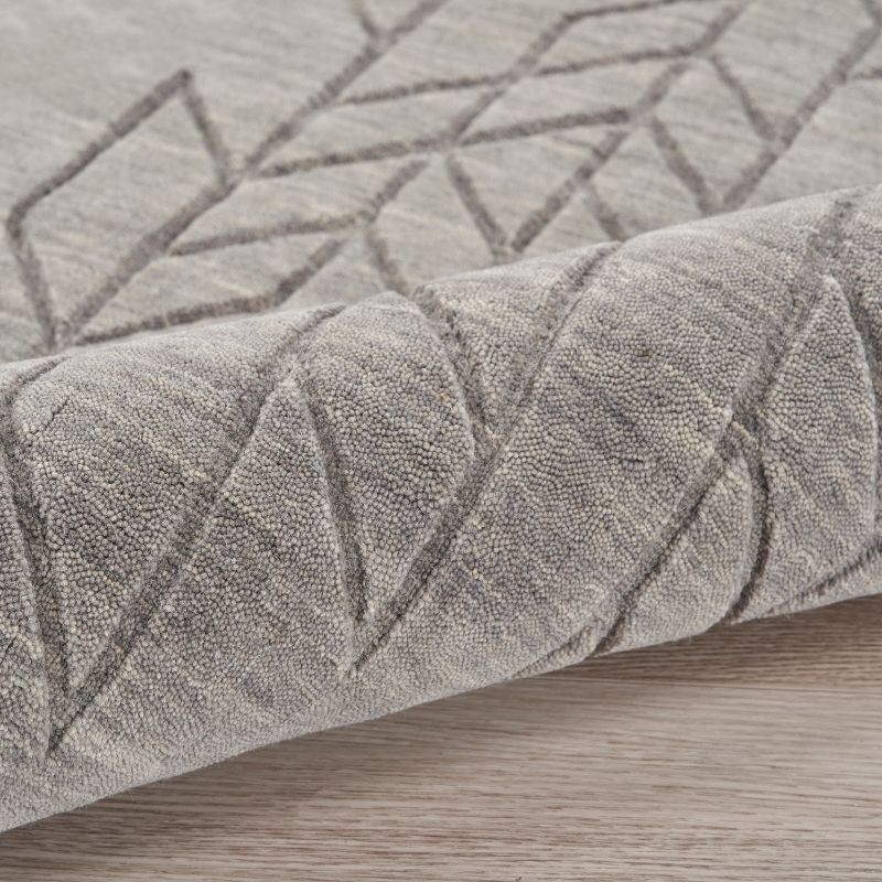 A close-up of a textured area rug in a warm gray or taupe color, featuring a raised geometric diamond pattern, shown folded over to reveal its pile and texture on a light wood floor.