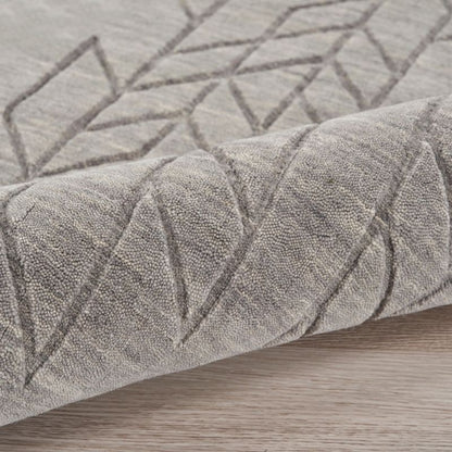 A close-up of a textured area rug in a warm gray or taupe color, featuring a raised geometric diamond pattern, shown folded over to reveal its pile and texture on a light wood floor.