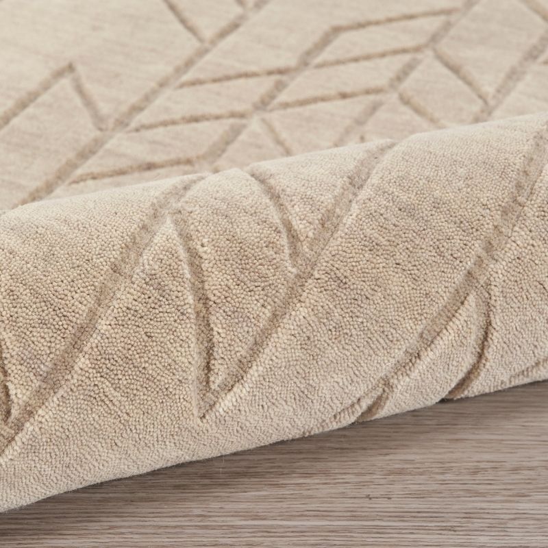 A close-up of a beige textured rug with a raised geometric leaf pattern, shown partially rolled over to reveal its pile and texture on a light wood floor.