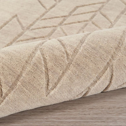 A close-up of a beige textured rug with a raised geometric leaf pattern, shown partially rolled over to reveal its pile and texture on a light wood floor.