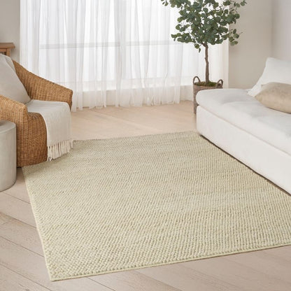 A large rectangular woven area rug in a light cream or off-white color, shown on a light wood floor in a bright, modern living room setting with a white sofa, a wicker armchair, and a potted tree.