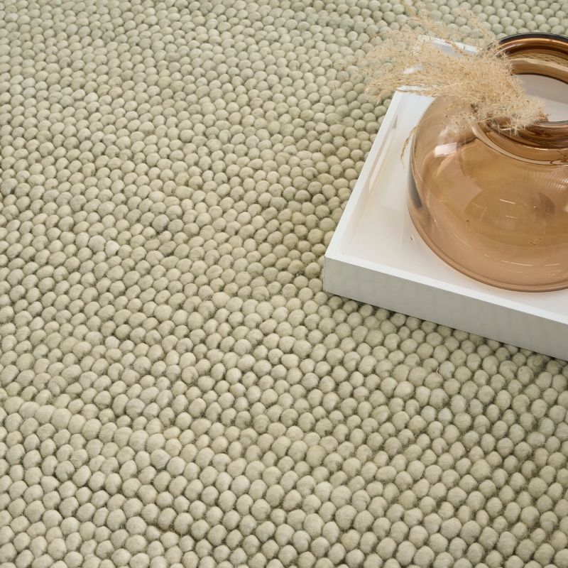 A close-up of a textured area rug with a dense, small-bobble pattern in a light beige or oatmeal color. A white rectangular tray holding a translucent amber glass vase with dried grass sits on the right side of the rug.