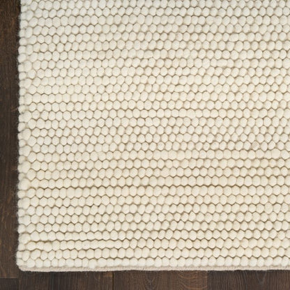A close-up of a cream-colored textured area rug with a raised honeycomb pattern, shown on a dark brown wooden floor.