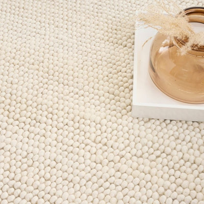 A close-up of a cream-colored, textured area rug with a dense tufted pattern, styled with a white block and a glass vase in the upper right corner.