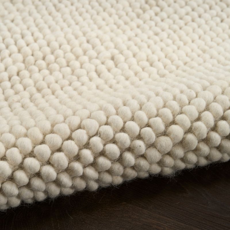 A close-up of a cream-colored wool area rug with a textured, raised tufted pattern, shown folded over to reveal its depth and texture against a dark wood surface.