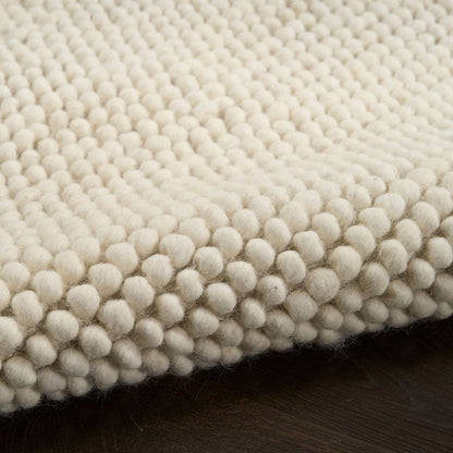 A close-up of a cream-colored wool area rug with a textured, raised tufted pattern, shown folded over to reveal its depth and texture against a dark wood surface.